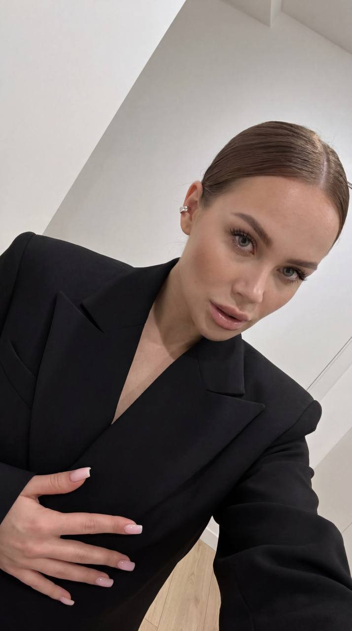 A low angle selfie of a woman in a black blazer, looking down into the camera. Her sleek chestnut hair and smooth features are highlighted.