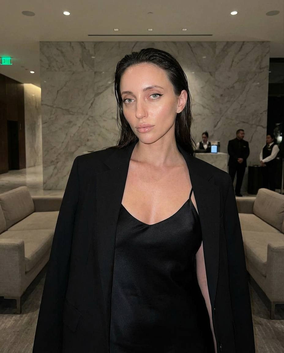 A cinematic photo featuring a model in a silk slip dress and oversized blazer, standing in a luxury hotel lobby with marble walls and blurred night staff.