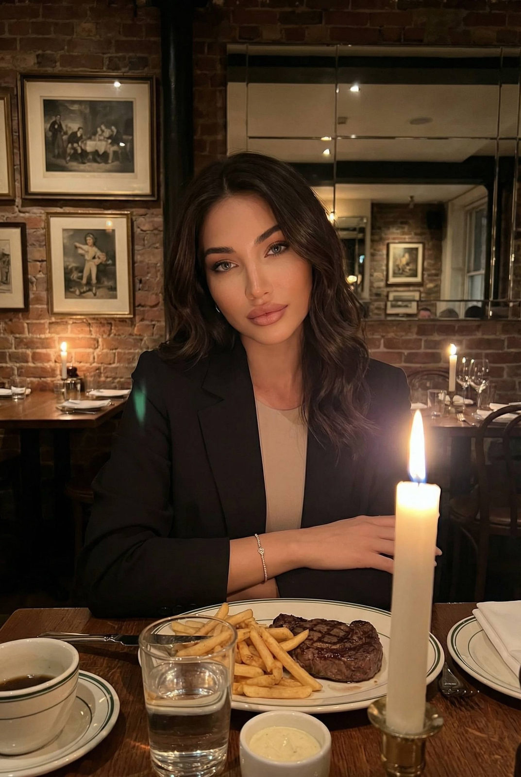 A woman in a tailored black blazer seated at a restaurant table, gazing calmly towards the camera in warm candlelight.