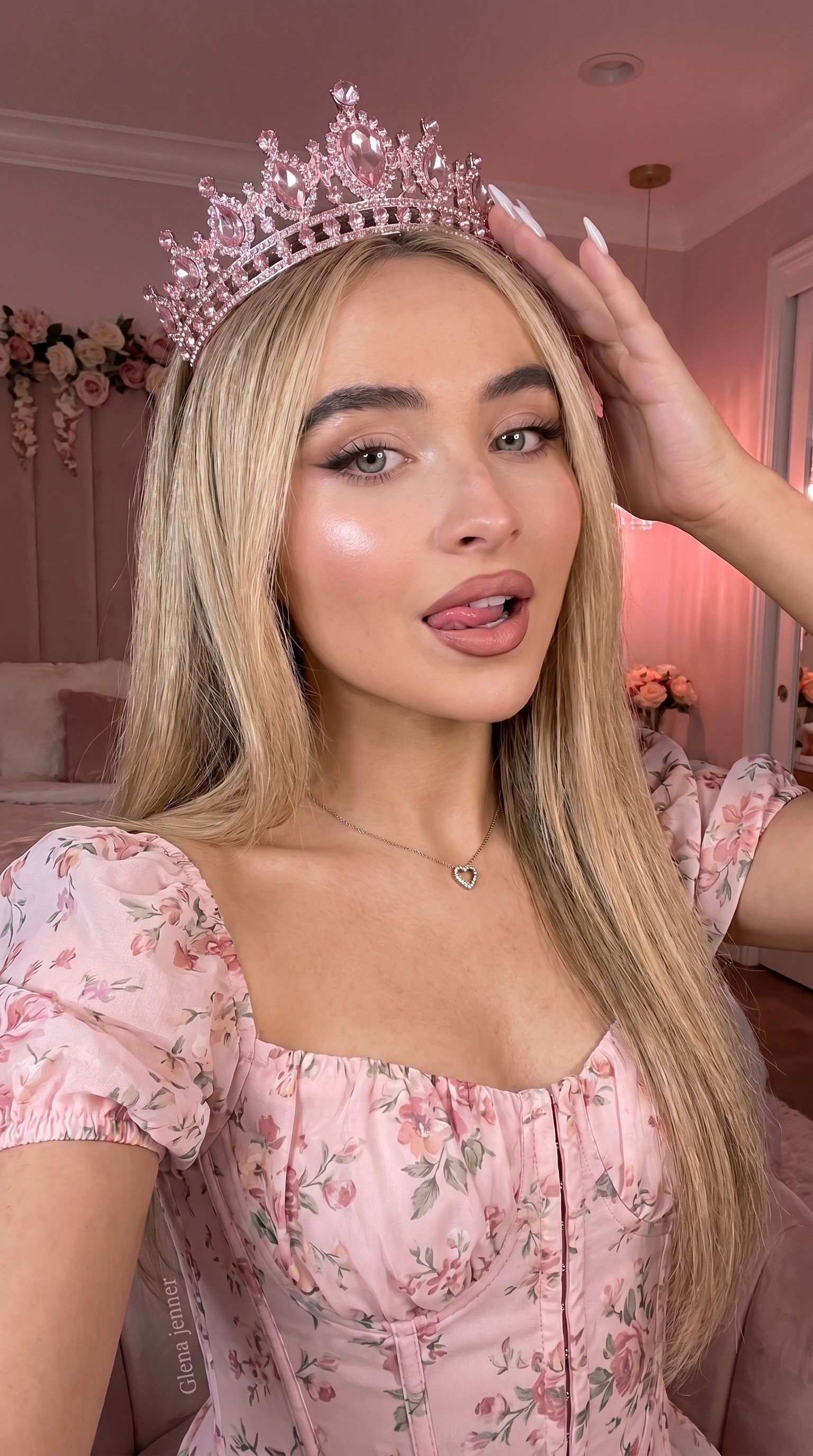A close-up selfie of a person wearing a sparkling pink tiara, light pink corset, and a playful expression in a soft pink bedroom.