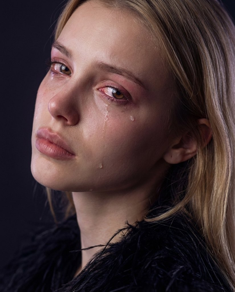 Dramatic close-up portrait of a girl with emotional expression, tear-streaked cheeks, and glossy eyes in a dark feathered garment.