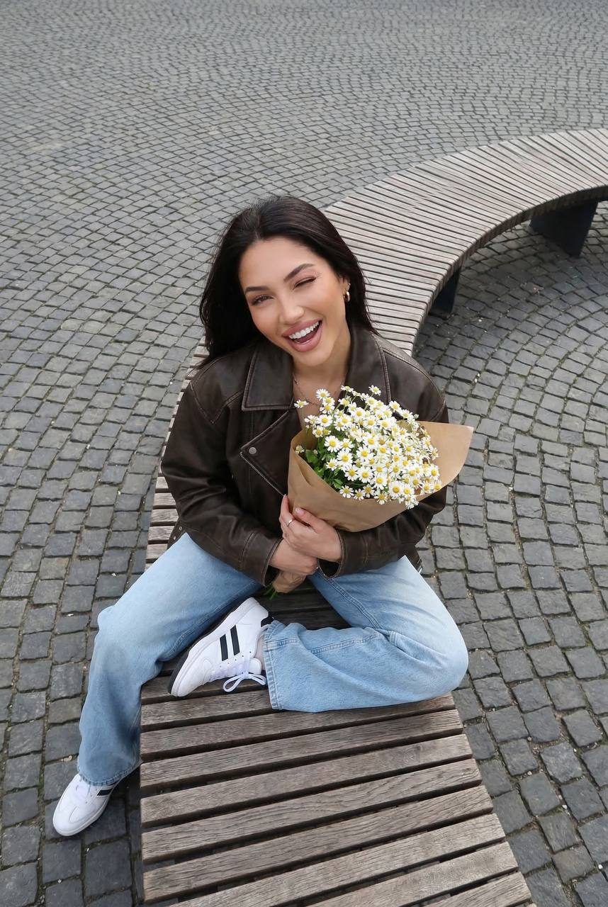 High-angle shot of a woman sitting on a curved wooden bench, holding a bouquet of daisies in an urban square.