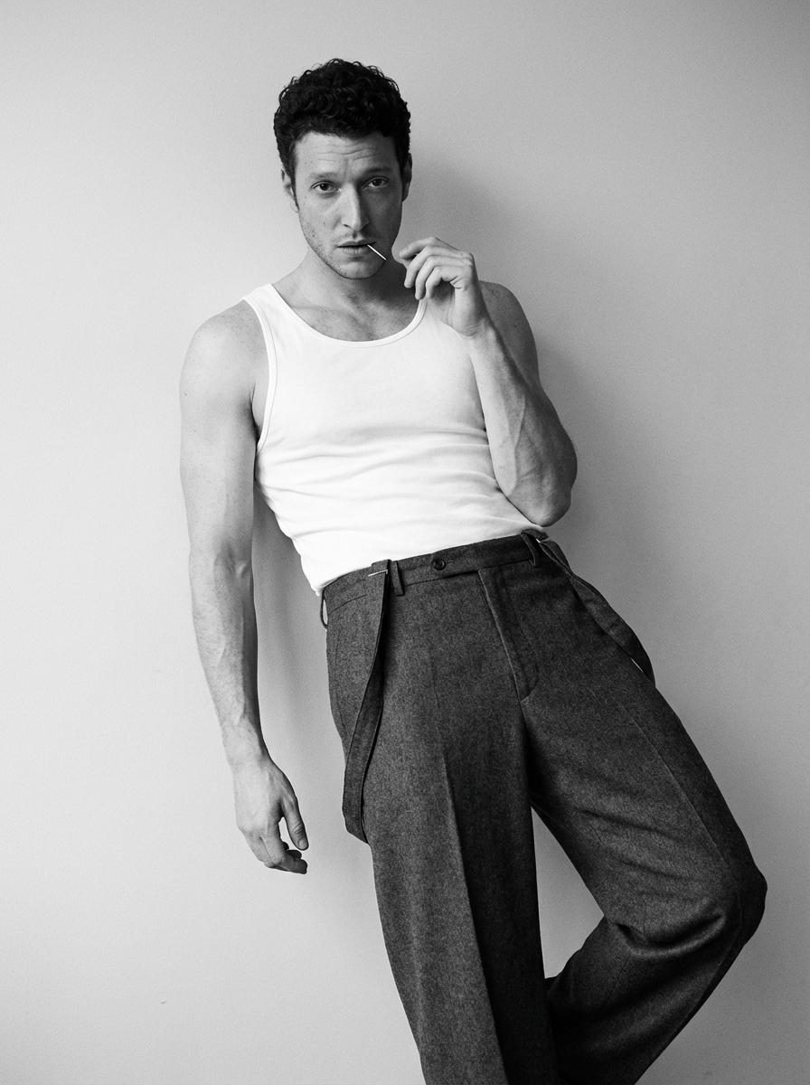 A man leaning against a light gray wall in a white tank top with a toothpick in his mouth, showcasing a relaxed S-curve pose.