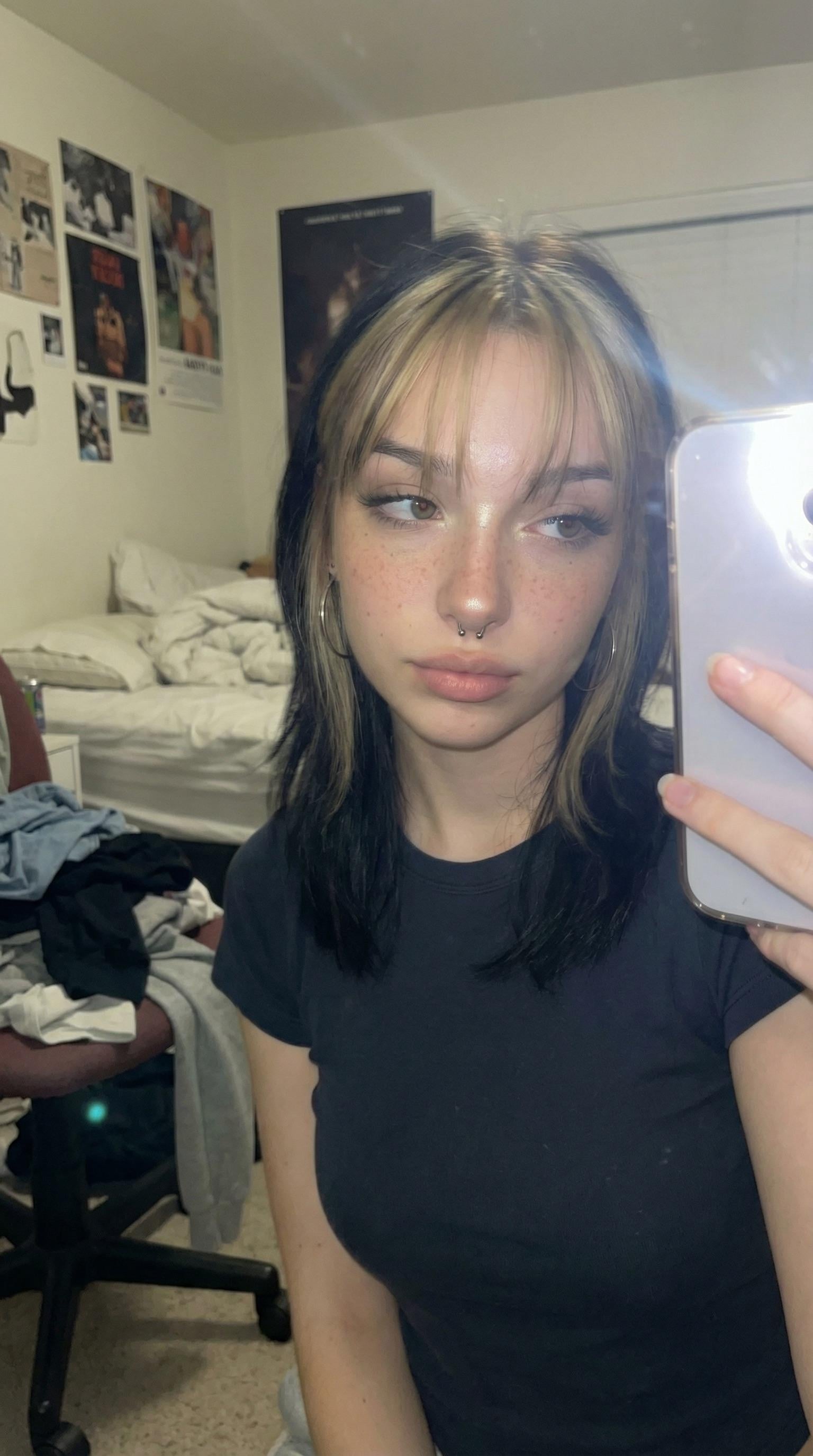 A close-up selfie in a bedroom, featuring a person in a black t-shirt with dark highlighted hair and hoop earrings, soft lighting enhances the scene.