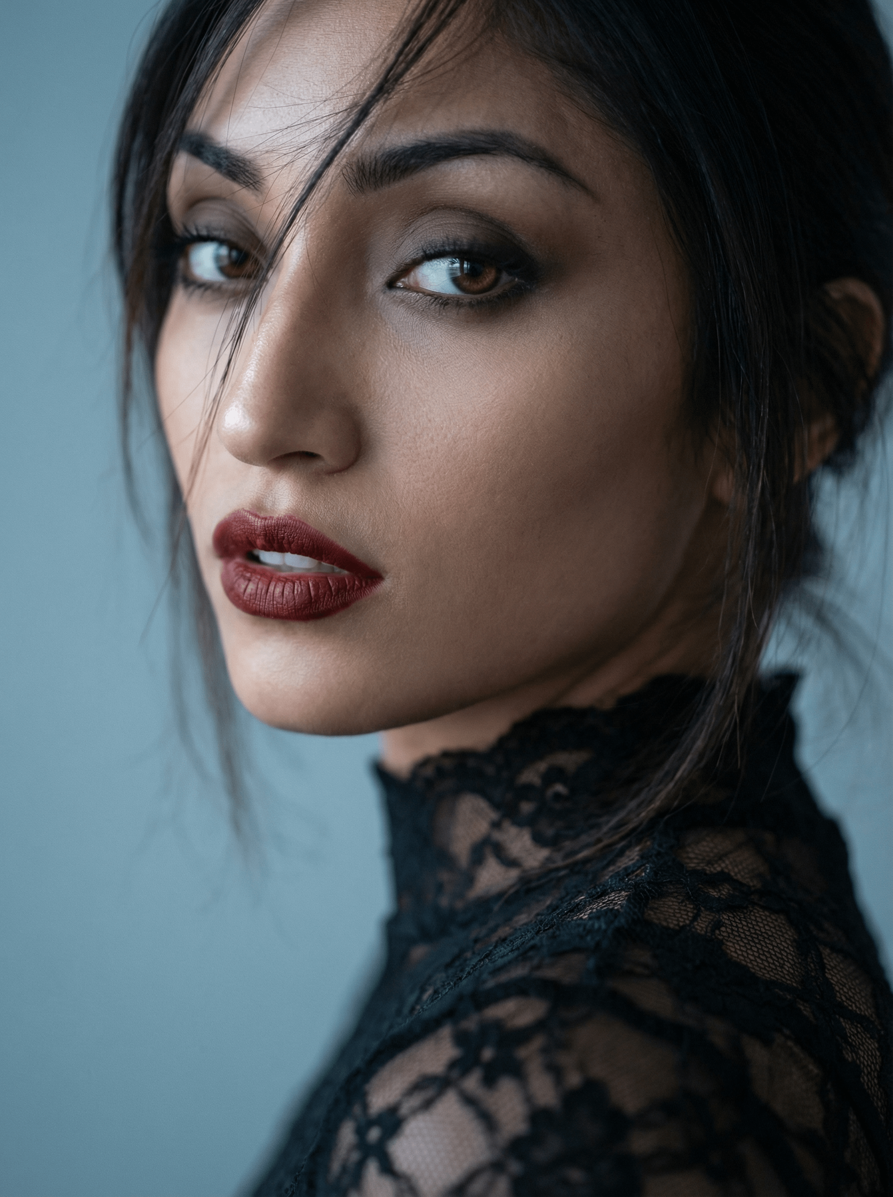 A close-up portrait of a gothic beauty with oxblood lips, soft blue-gray background, and gentle lighting accentuating facial features.