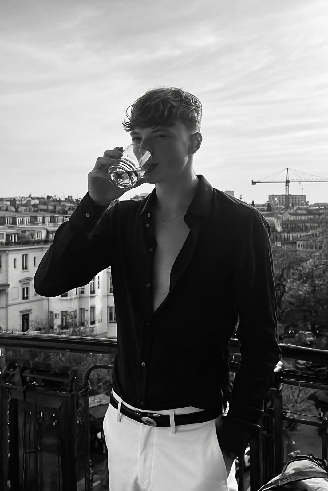 A man on a balcony, relaxed, holding a glass, surrounded by urban buildings with a soft monochrome tone.