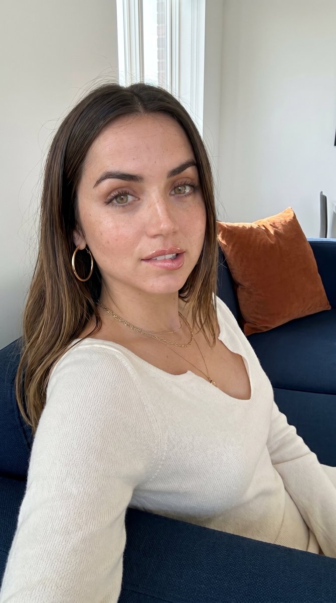 Ultra-realistic close-up portrait of a woman with long hair on a modern sofa, wearing gold hoop earrings and a knit top with a soft expression.