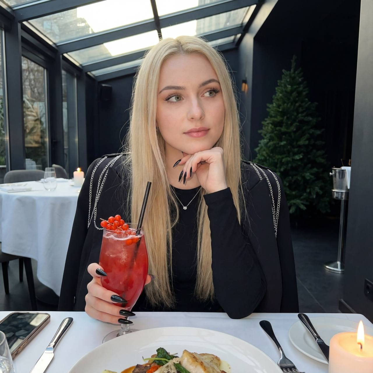 A woman sitting at a table in a modern restaurant, wearing a blazer and holding a red cocktail, with a green Christmas tree in the background.