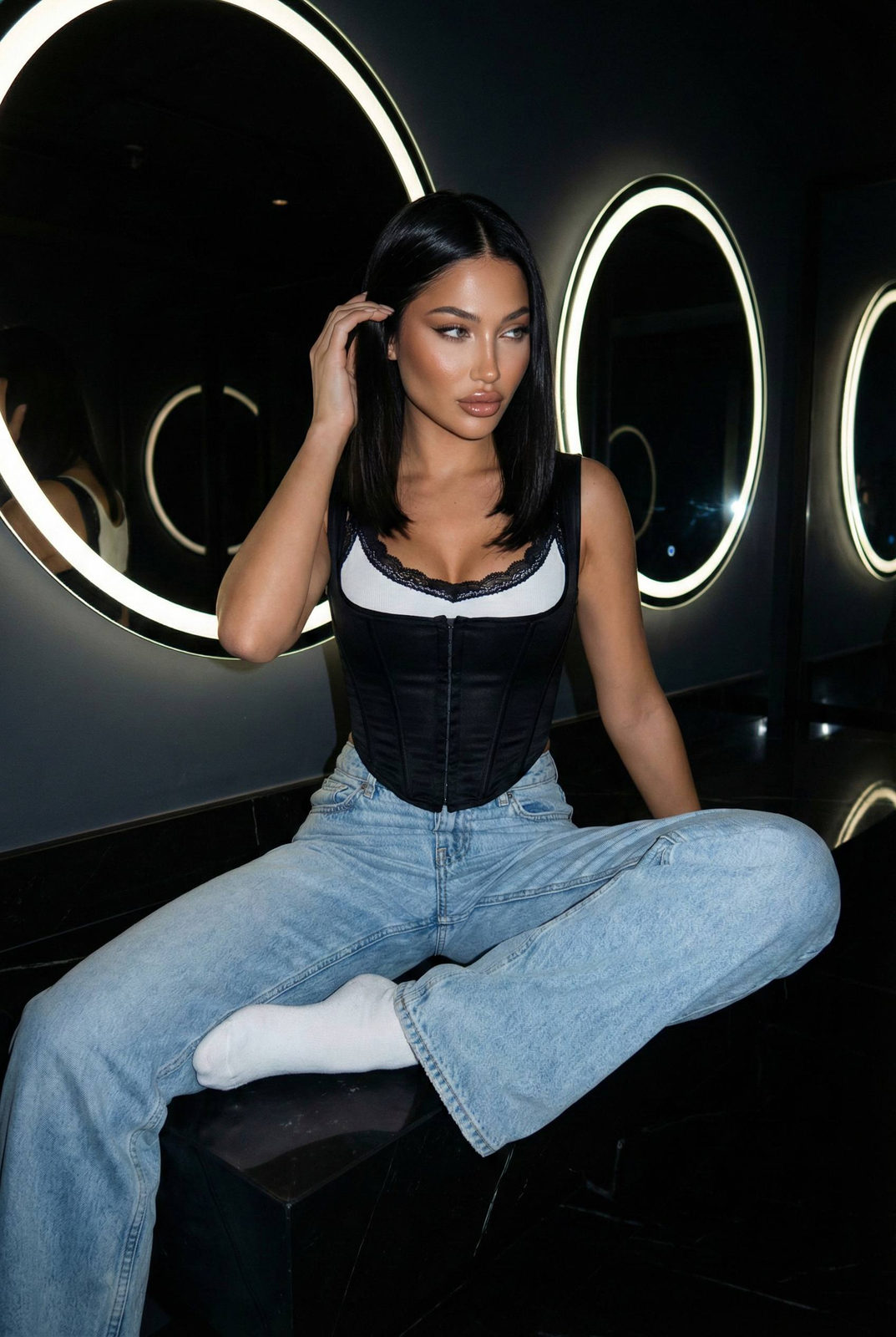 Woman sitting relaxed in a modern lounge with circular mirrors, dressed in light-wash jeans and a corset.