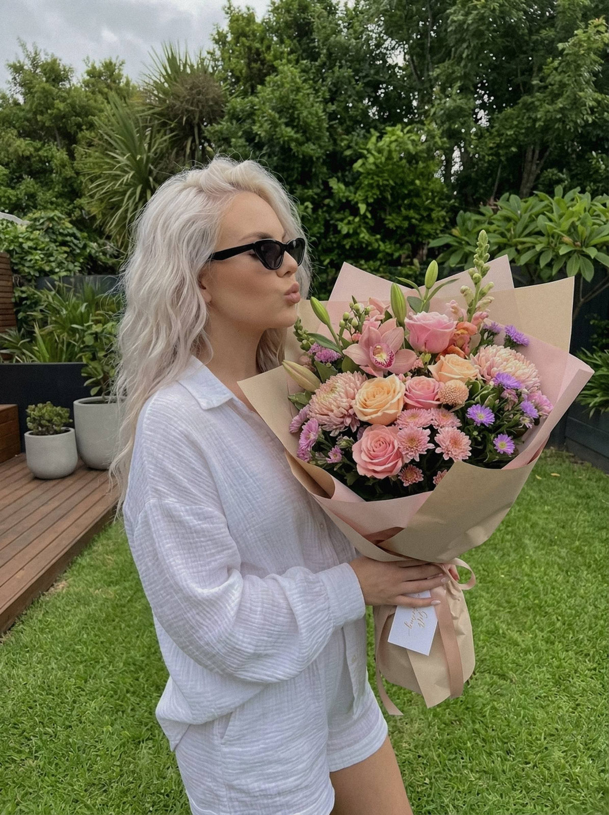 A woman in a garden, gently kissing a large bouquet, showcasing Y2K glam makeup and a relaxed summer outfit.
