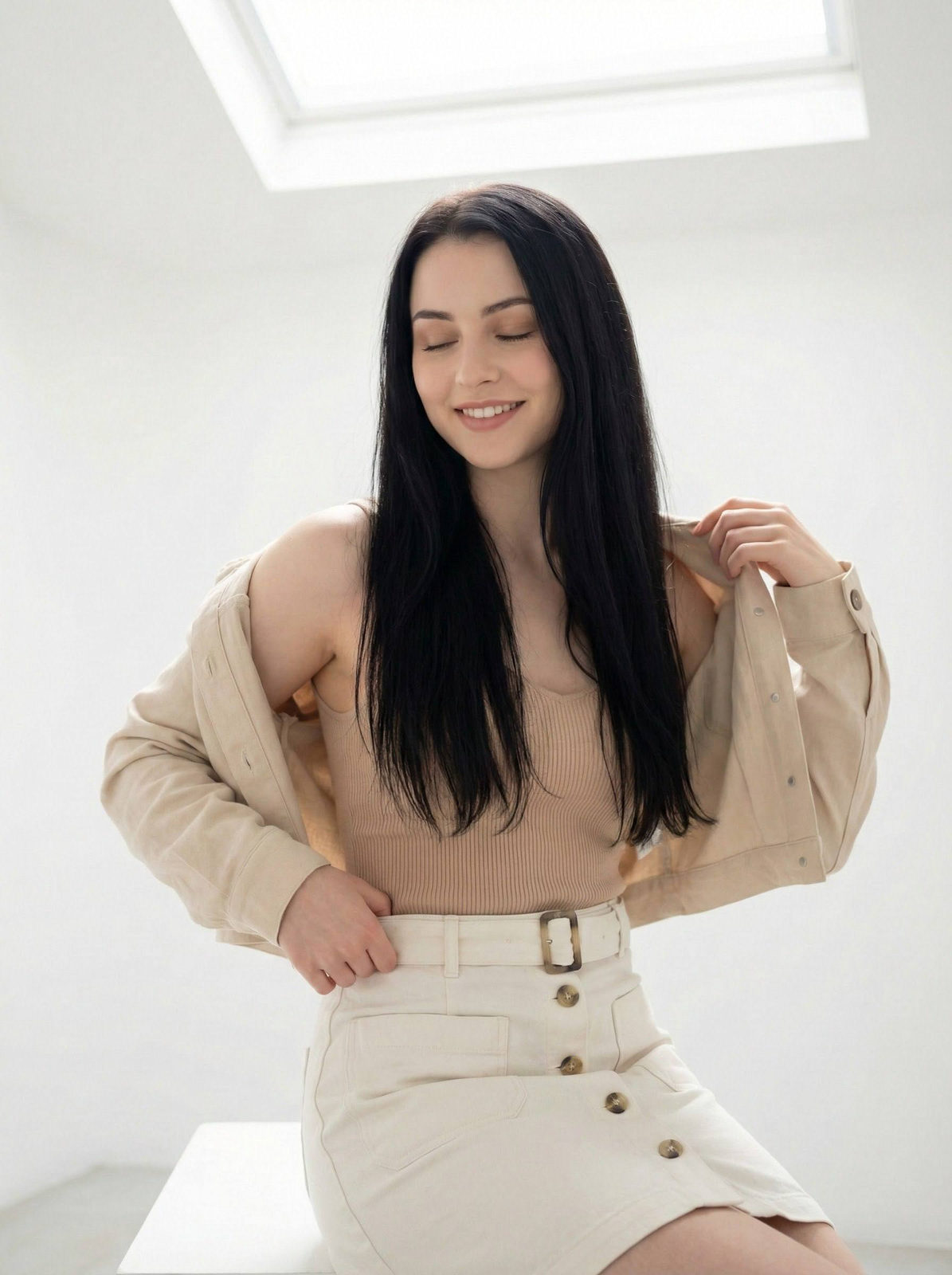 A woman in a bright minimalist white studio, lifting her jacket off her shoulder, with long straight hair and a gentle smile.