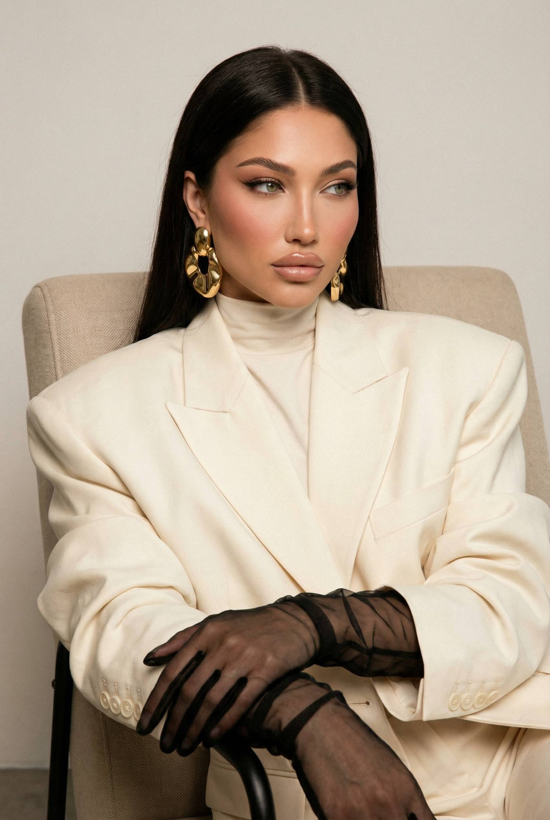 Close-up portrait of a woman in an ivory blazer seated in a beige armchair, showcasing luminous skin and sculpted features.