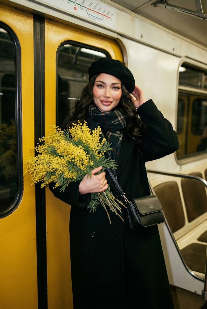 A woman in a black coat and beret stands near vintage subway doors holding a bouquet of mimosa flowers.