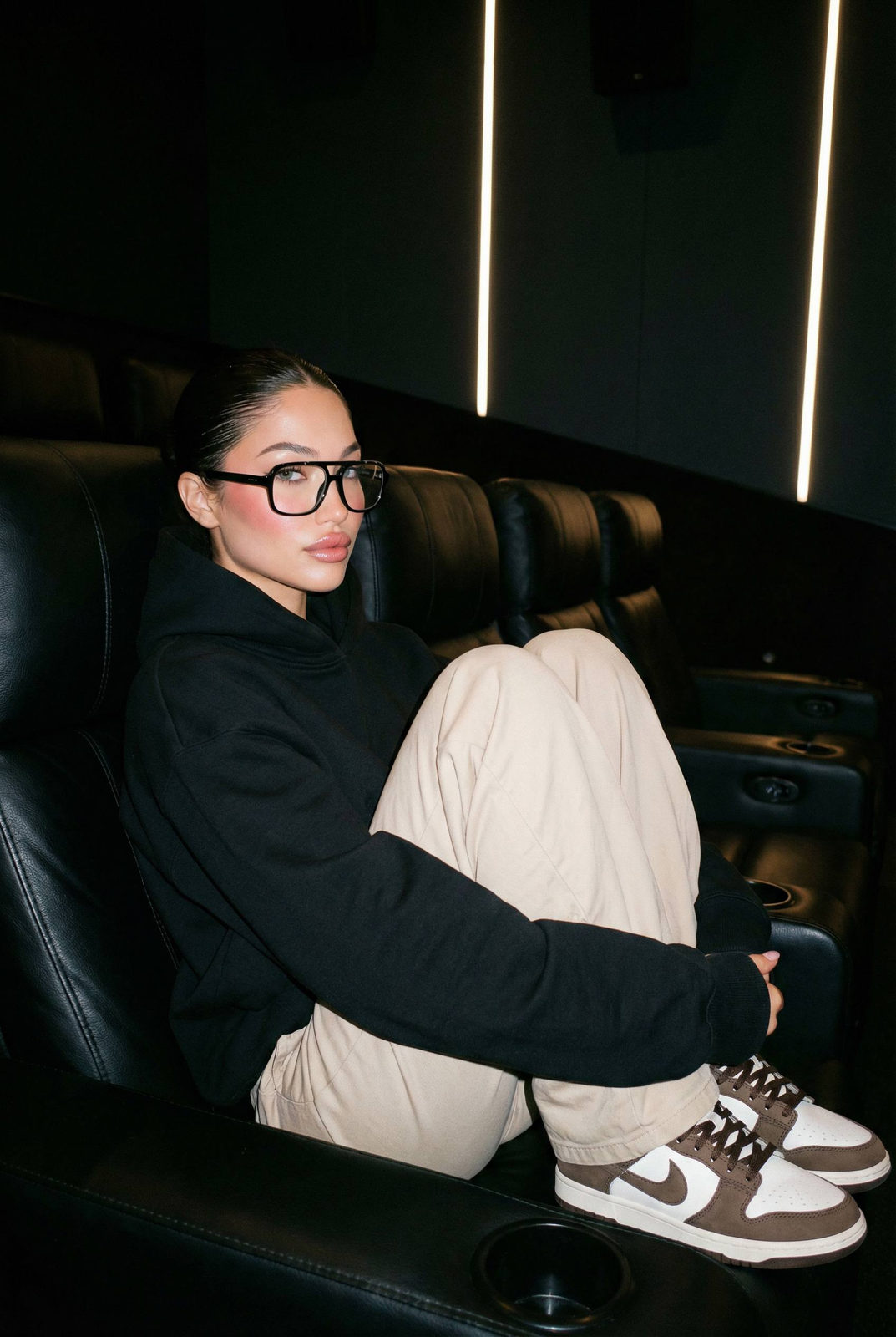 A cozy woman in a black hoodie, sitting curled up in a modern cinema chair, surrounded by dark acoustic walls and soft LED lighting.
