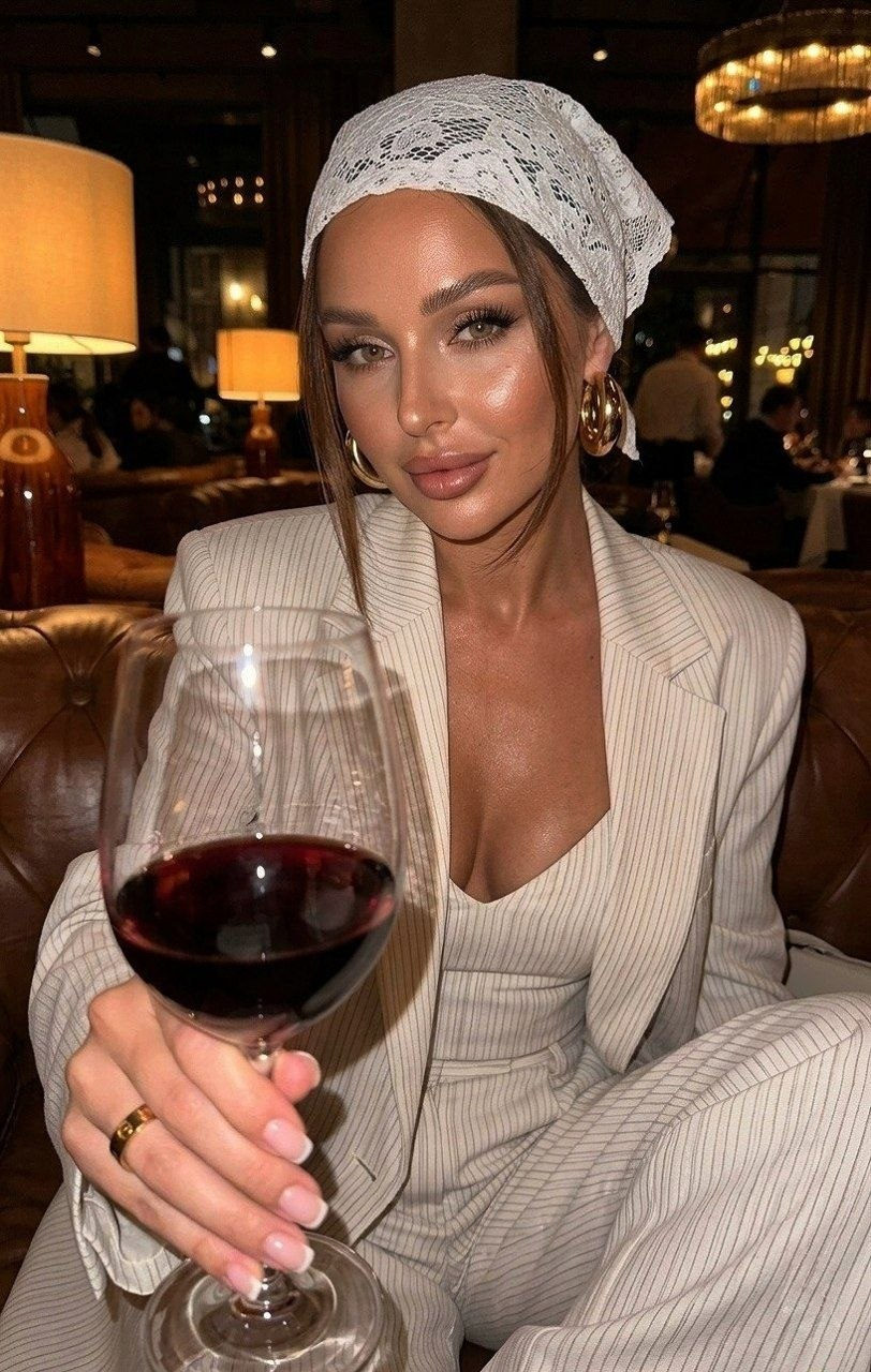 Photorealistic selfie of a woman in a pinstriped suit holding a glass of wine, soft makeup, glowing skin, warm lighting.