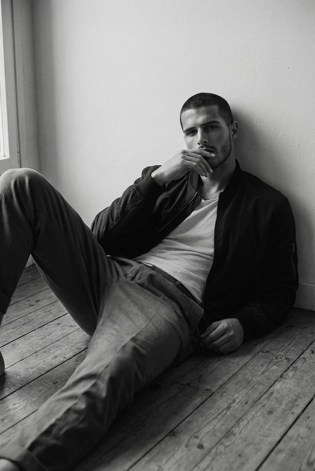 Ultra-realistic black and white portrait of a young man reclining on a wooden floor, gazing contemplatively into the camera.