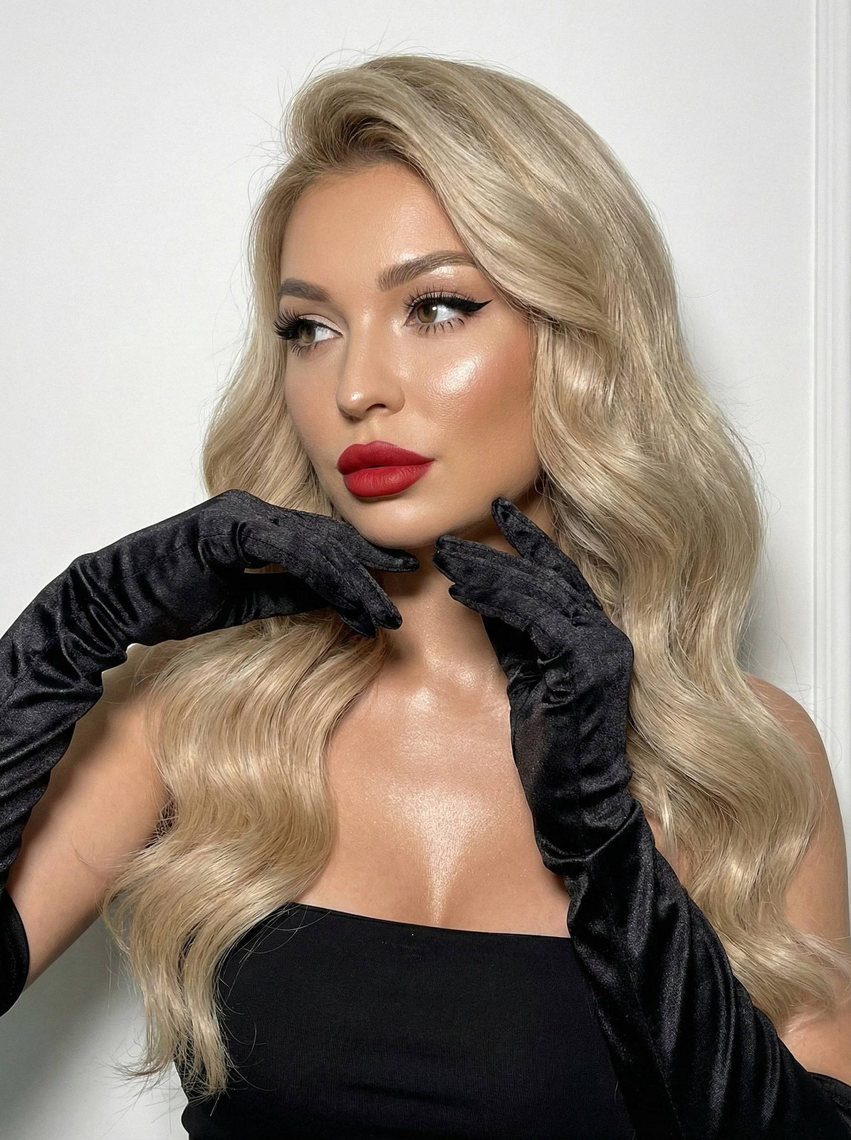A close-up of a woman in a strapless top and satin gloves, looking thoughtfully to the side with dramatic lighting enhancing her flawless makeup and glowing skin.