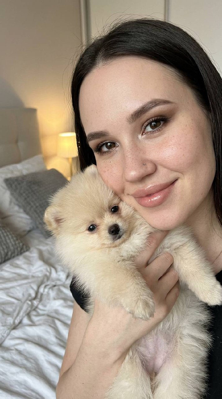 A young girl with freckled skin smiling, holding a cream-colored Pomeranian puppy in a softly lit bedroom.