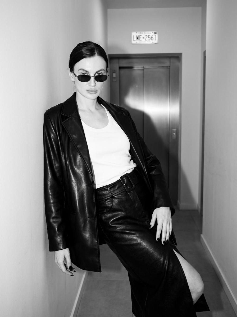 A woman in a narrow apartment hallway wearing a black leather jacket, leaning against the wall, captured in high-contrast monochrome.