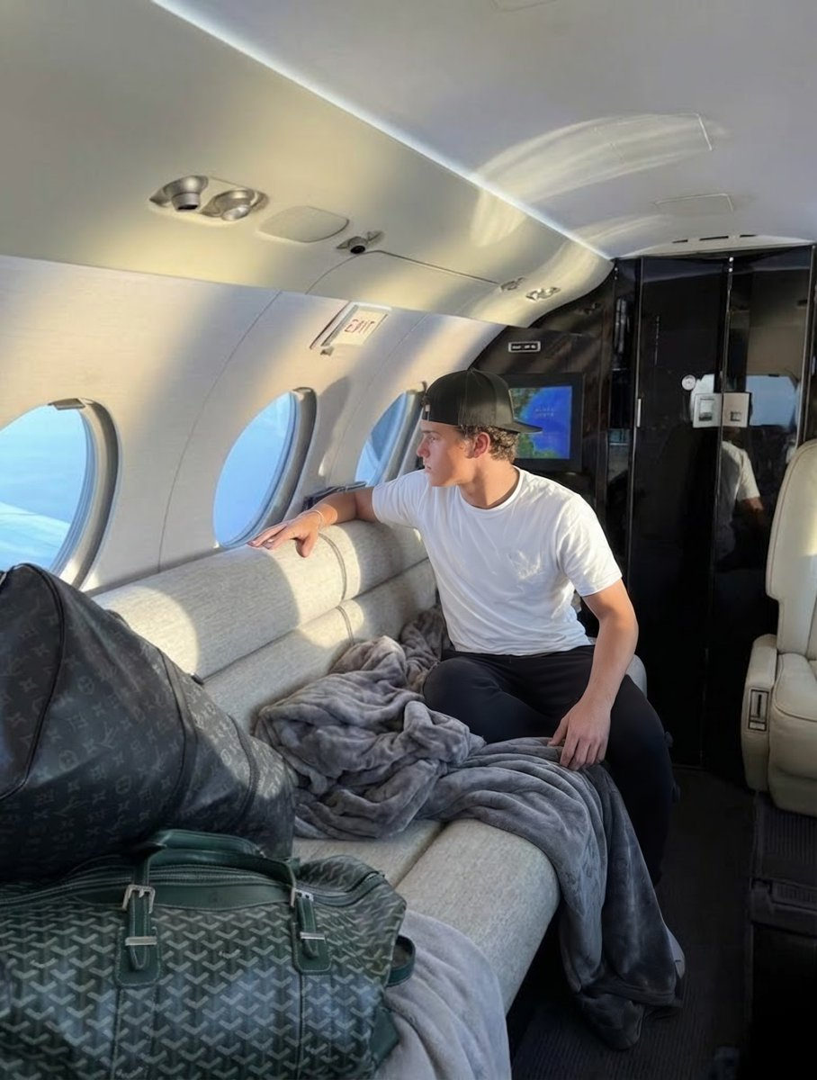 A young man in a casual pose seated in a luxurious jet cabin, gazing out the oval window surrounded by soft gray blankets and a travel bag.
