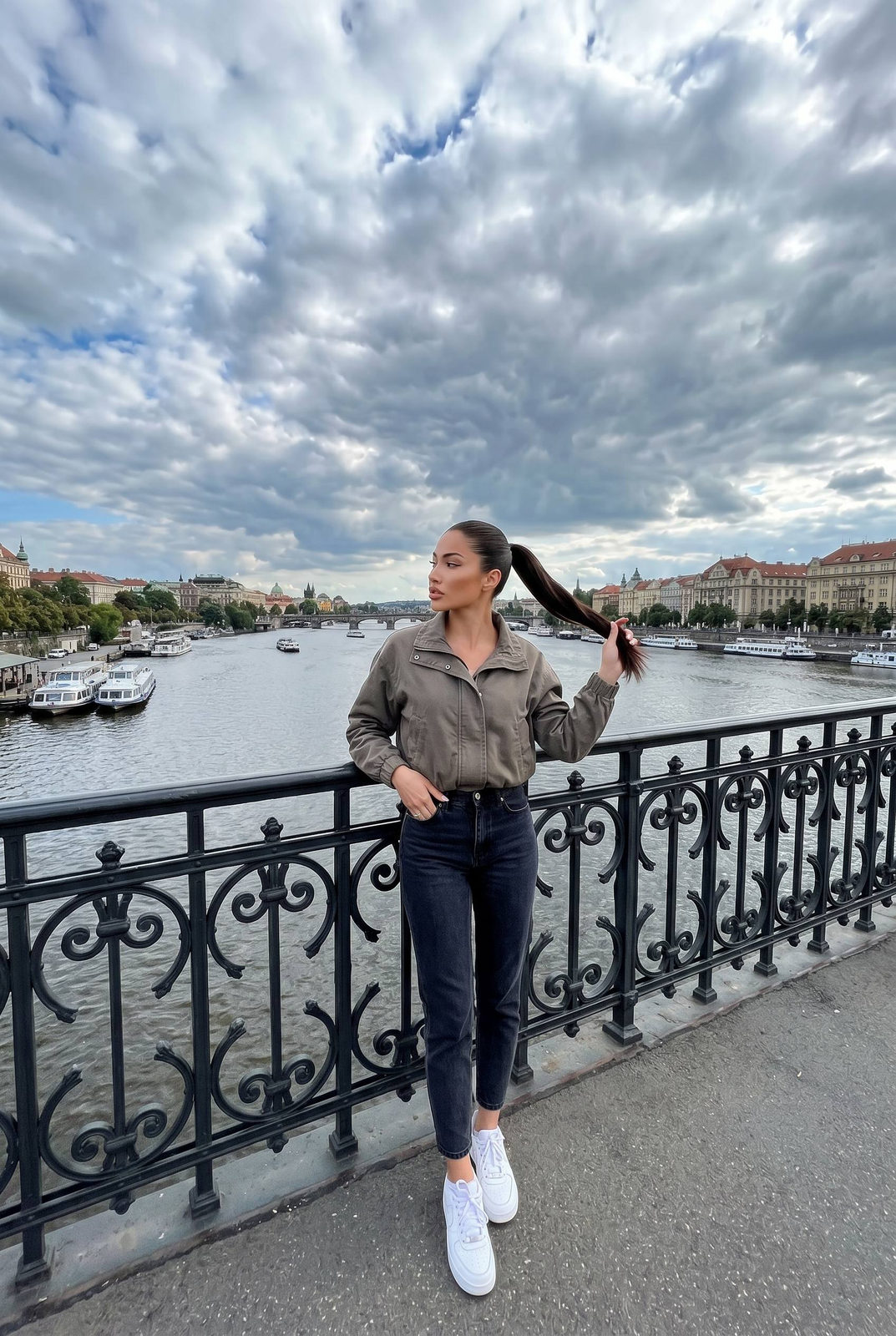 A woman casually stands on a historic bridge, overlooking a river with dramatic clouds, creating a cinematic atmosphere.