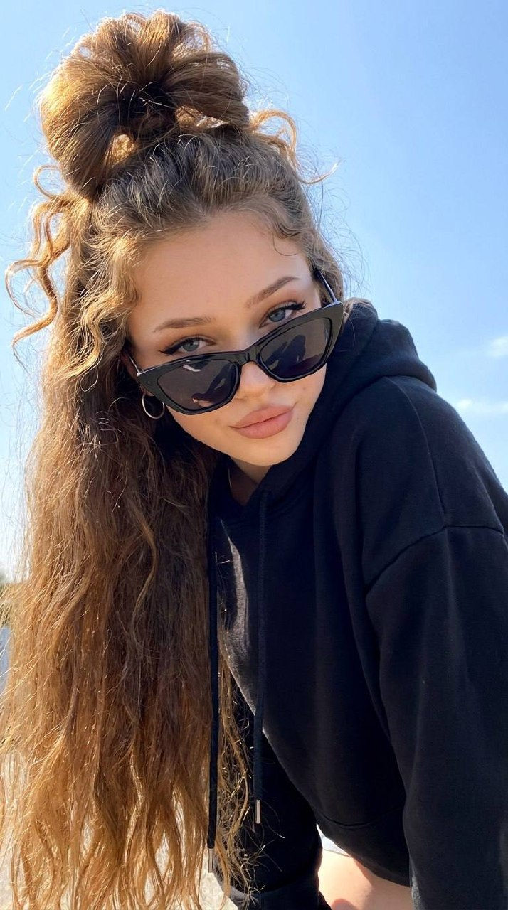 A girl with a messy bun and sunglasses leaning forward in bright sunlight, wearing a black hoodie against a blue sky.