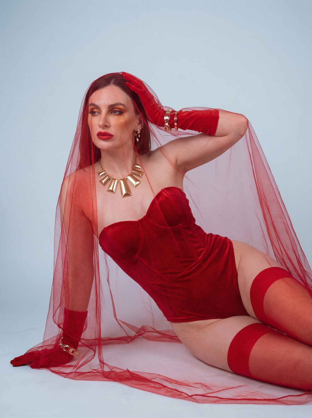 A woman reclining in a studio, wearing a crimson corseted bodysuit with a flowing red veil, illuminated by soft lighting.
