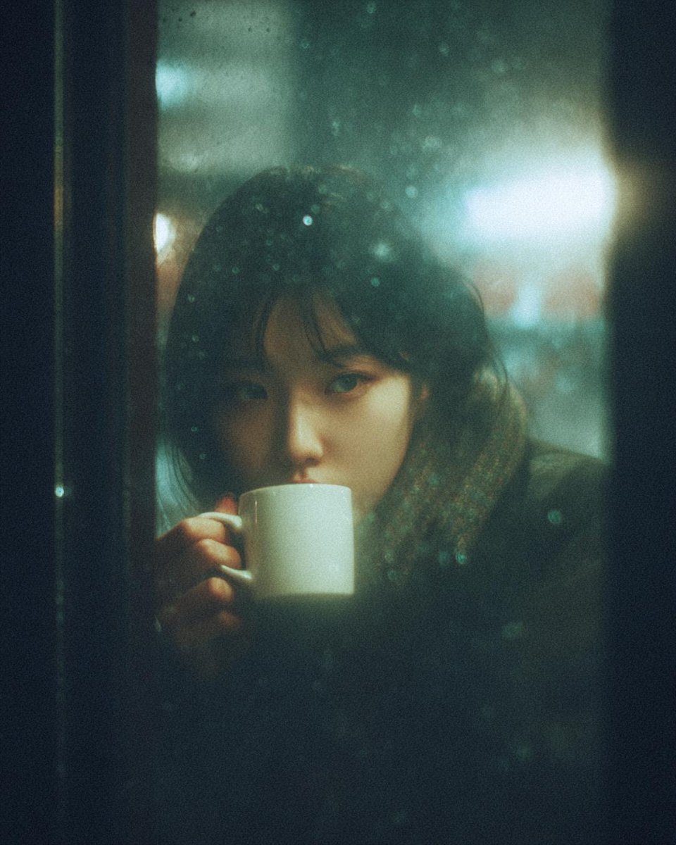 A woman with dark hair sitting at a café, viewed through a rain-covered window, holding a ceramic mug.