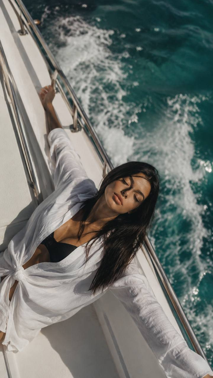 A person lying on a boat deck with arms spread, dressed in a white translucent shirt and dark swimsuit, beside turquoise water under natural light.