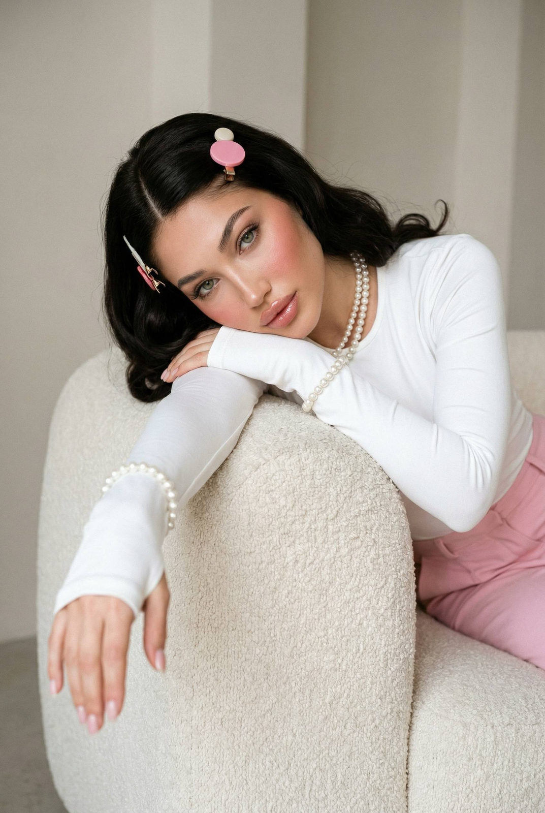 A woman in a white top and pastel pink skirt seated in a cream boucle armchair, leaning elegantly with glossy hair and pearl accessories.