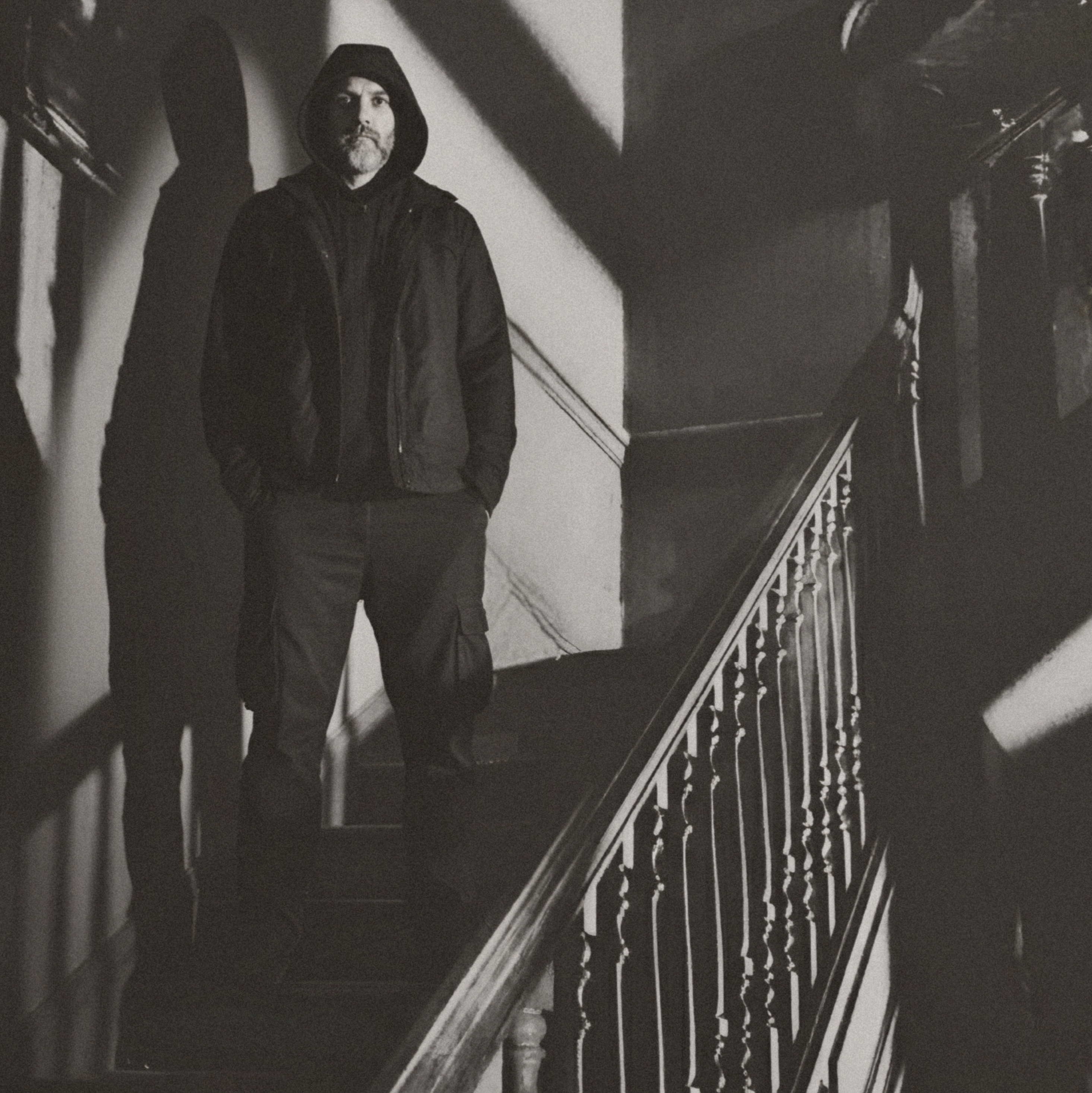 Cinematic black and white photo of a wooden staircase in a decaying house, highlighting shadows and a somber man in a dark hooded jacket.