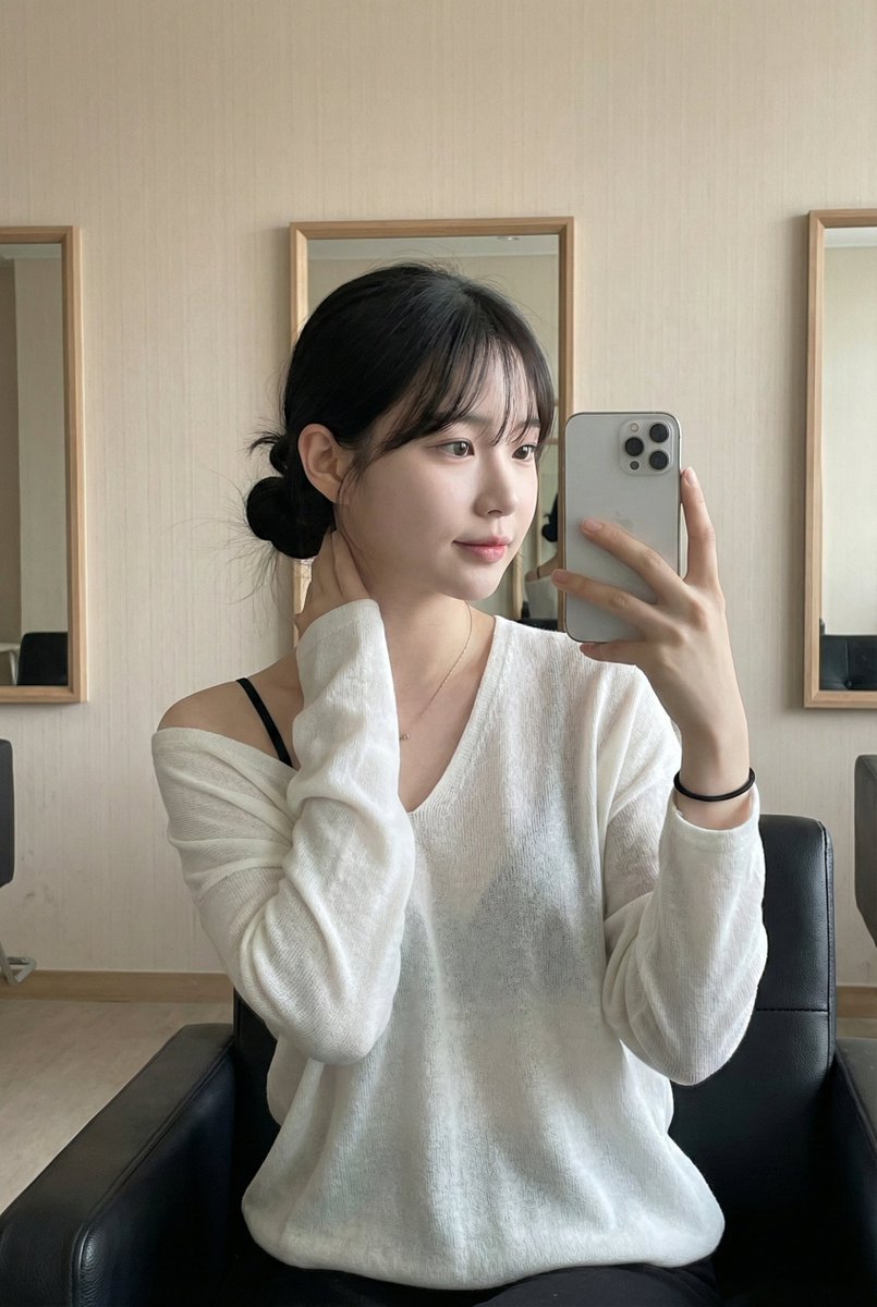 A woman in a leather chair taking a mirror selfie, showcasing a relaxed pose with a sleek ponytail and soft natural makeup.