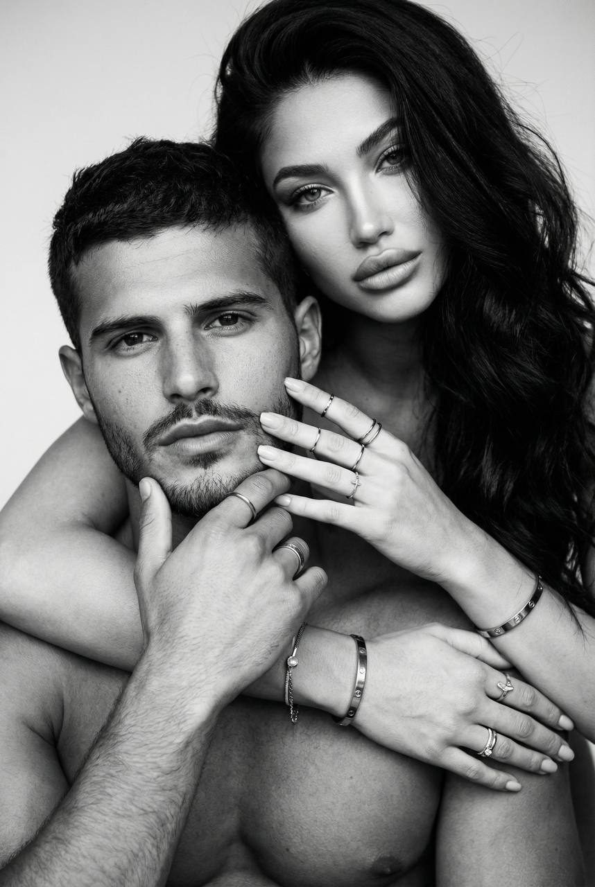 A close-up of a woman leaning over a man's shoulder, showcasing dramatic eye makeup and glossy lips in a soft high-contrast black and white style.