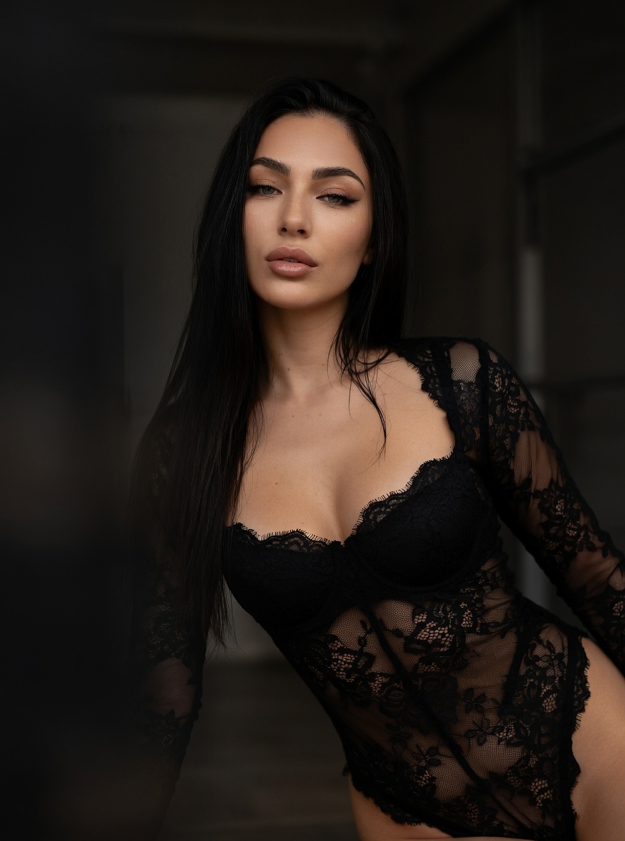 Low angle portrait of an East female model in a black lace bodysuit, softly lit with a warm amber color profile and a blurred dark background.