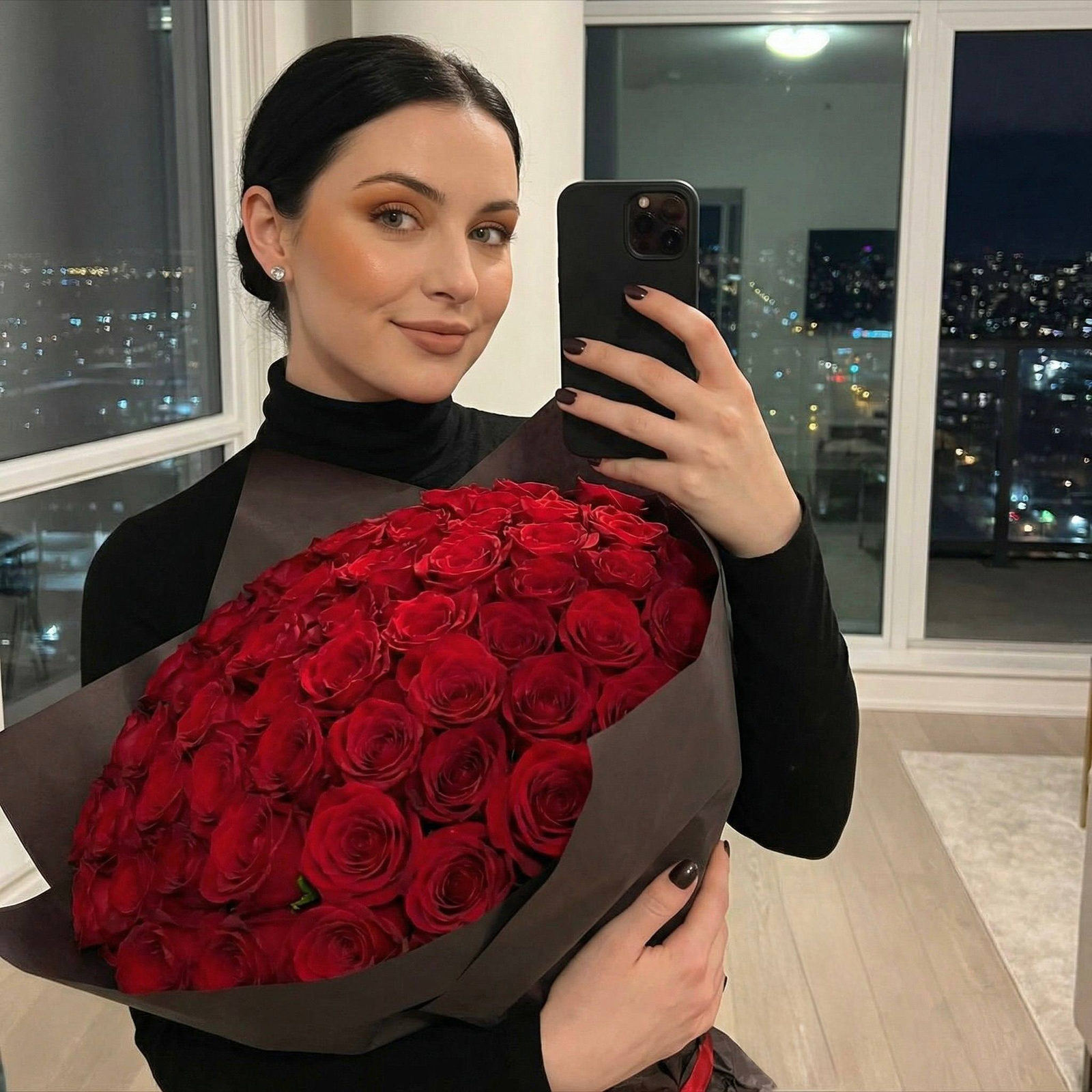 A woman in a fitted turtleneck poses with a large rose bouquet in front of a vibrant night cityscape, reflecting warmth and elegance.
