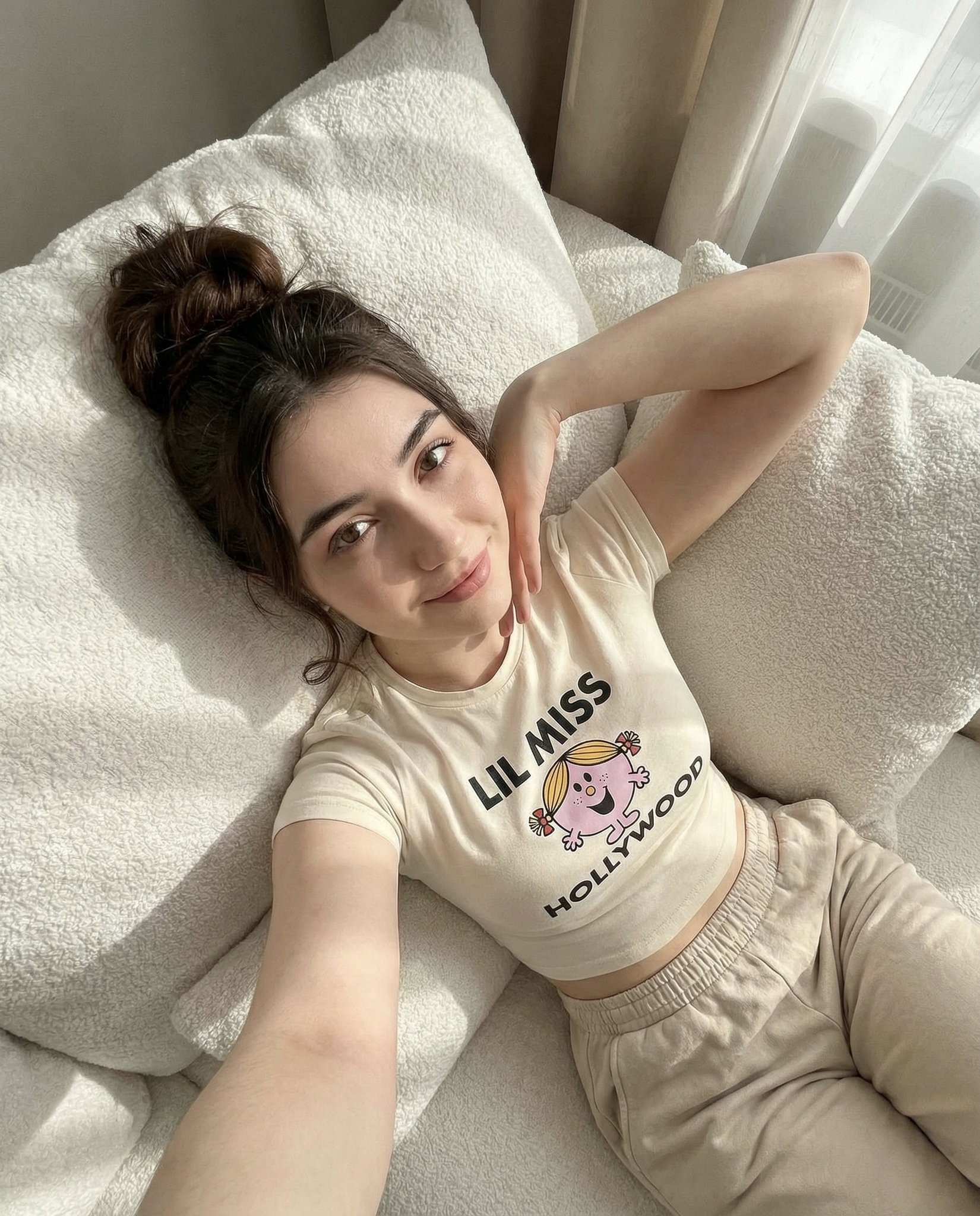 A woman in a cream white fitted cropped t-shirt and light beige casual bottoms, reclining on textured white pillows, gazing at the camera with a soft smile and messy bun hairstyle.