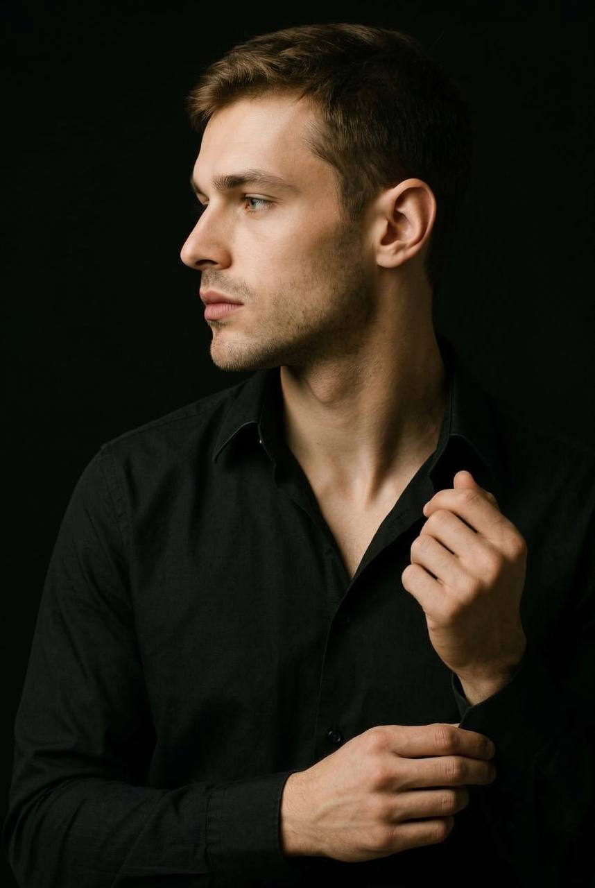 Dramatic dark studio portrait of a man in a black shirt against a deep black background, showcasing sculptural profile and soft lighting.