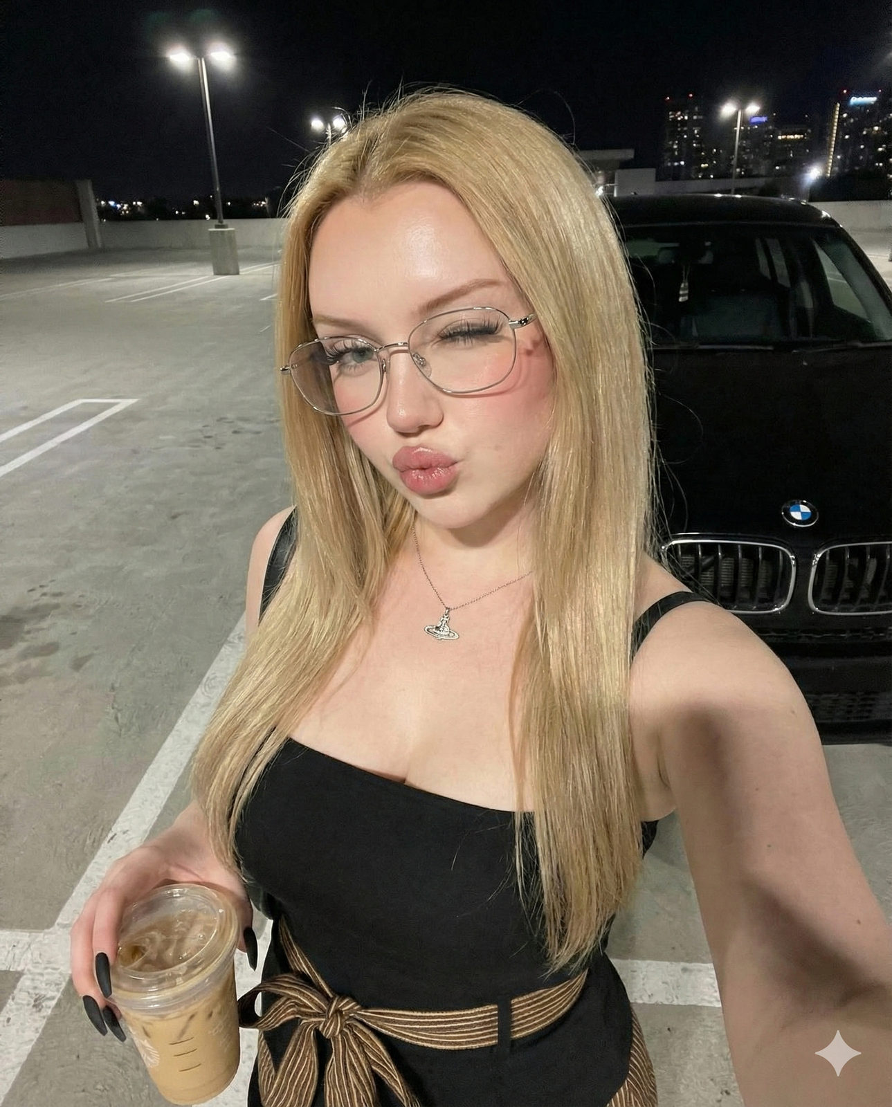 A young woman in a strapless top winking confidently while holding an iced coffee in a nighttime outdoor setting.