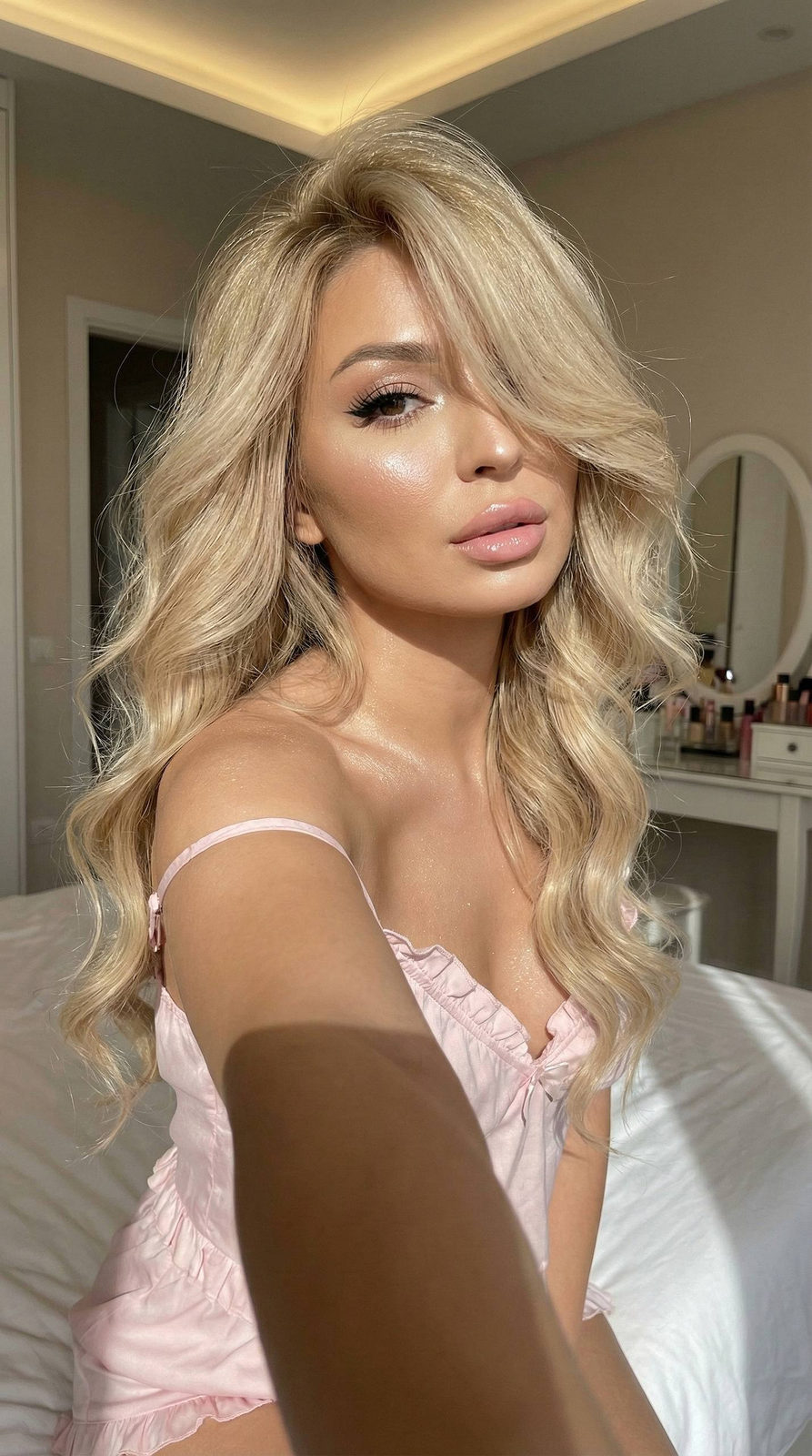 A close-up of a woman with voluminous hair, glowing skin, and a soft pink pajama in a sunlit bedroom.
