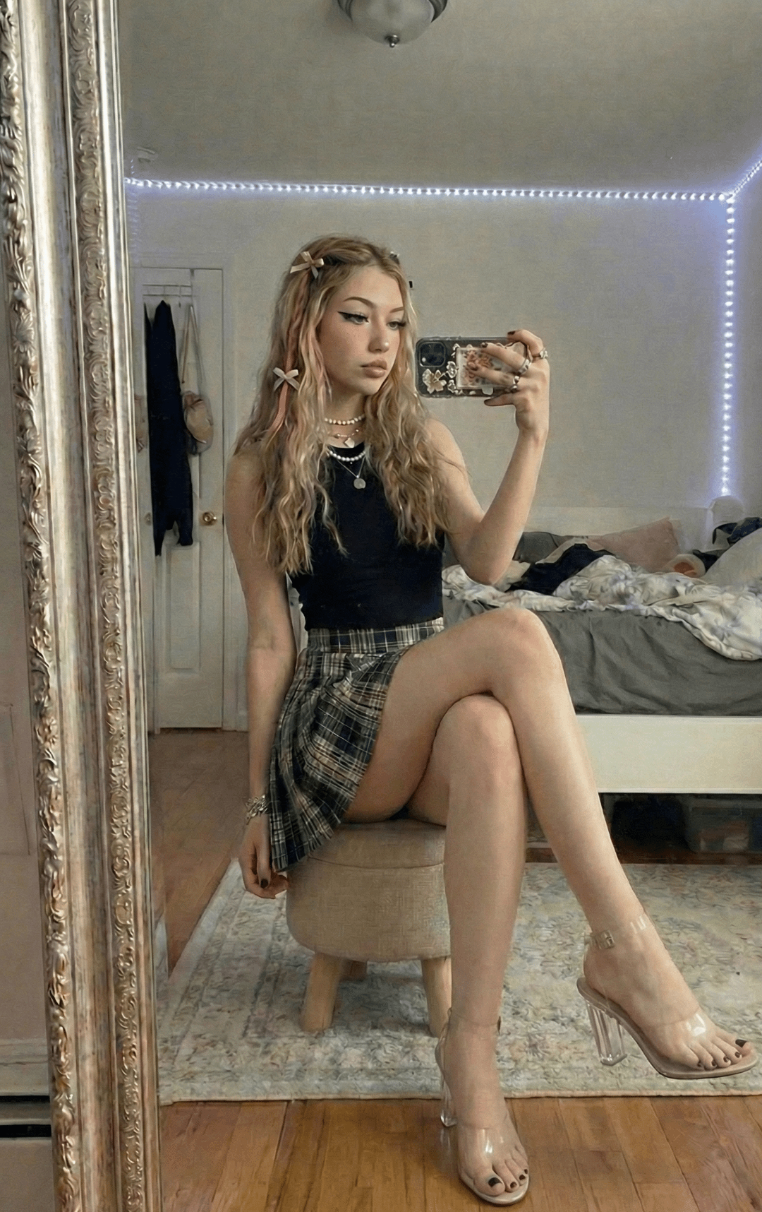 A photorealistic mirror selfie of a woman in a plaid skirt sitting on a beige stool, capturing her reflection in a spacious bedroom.