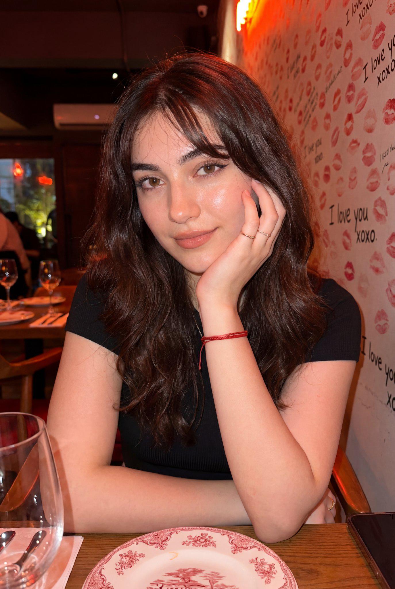 A woman in a black ribbed knit top seated at a wooden table in a dimly lit restaurant, leaning on her hand, with a backdrop of lipstick kiss marks and neon orange glow.