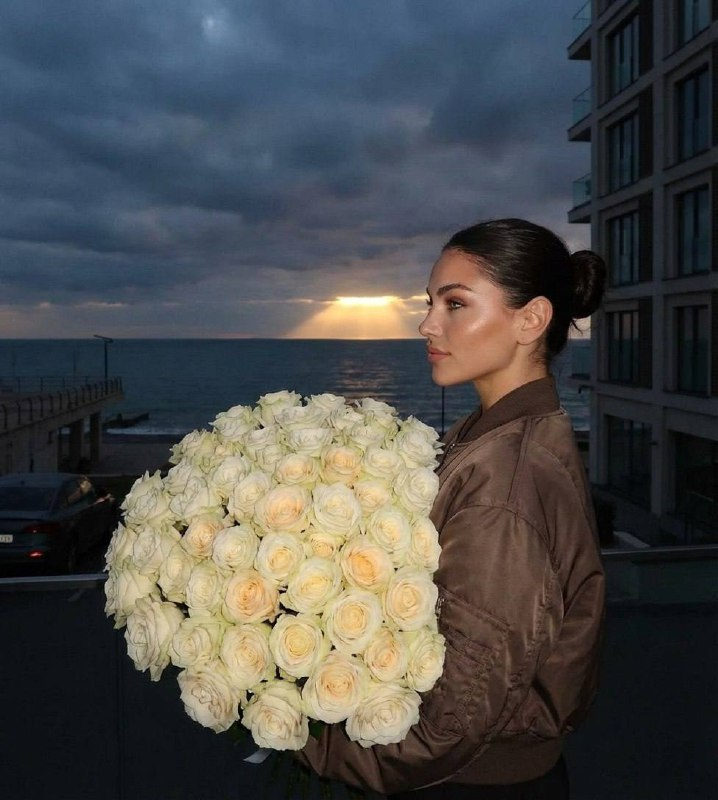 A profile shot of a girl in a brown bomber jacket holding a bouquet of cream roses against a twilight sea backdrop.