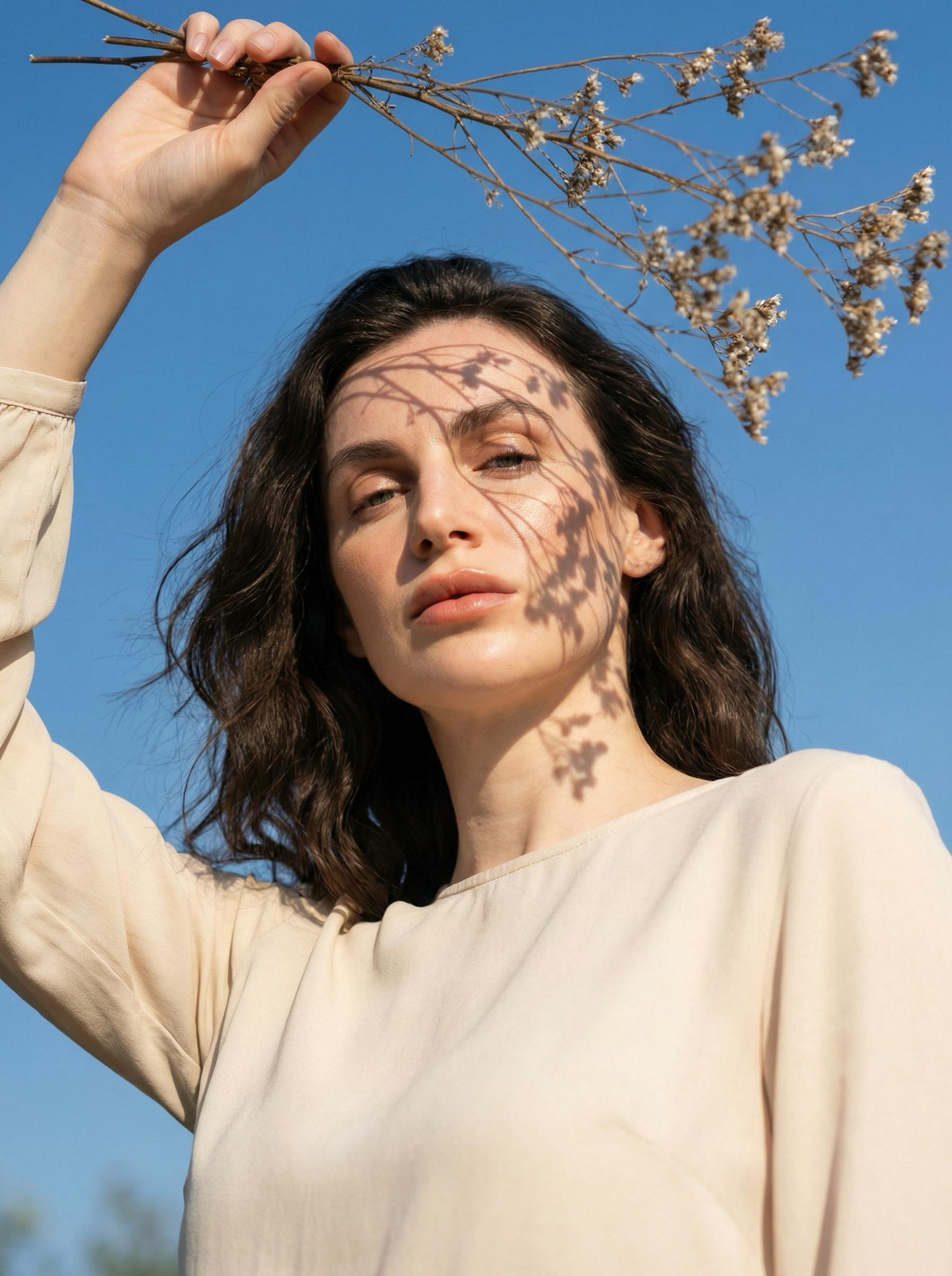 A woman photographed from a low angle, holding dried flowers above her head, creating intricate shadows on her face.