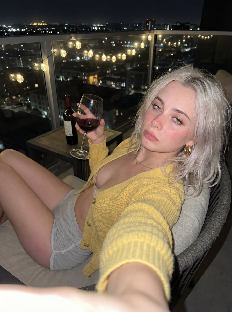 A woman with platinum blonde hair sitting on a cozy balcony chair, showing an inviting expression with city lights in the background.