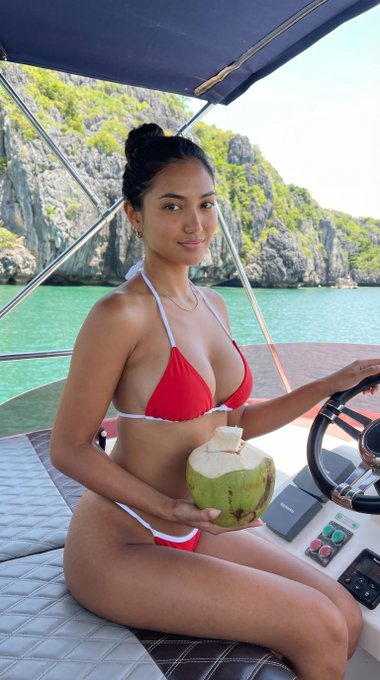 A woman in a red bikini seated at a boat helm, holding a green coconut, turquoise water and cliffs in the background.