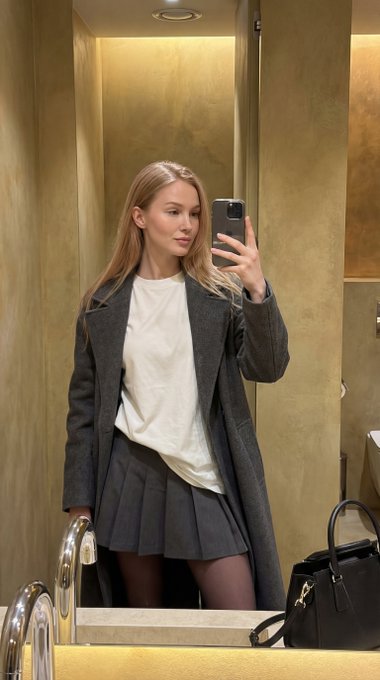 Woman taking a mirror selfie in a restroom with a golden wall and chrome faucet, dressed in a charcoal coat and dark mini-skirt.