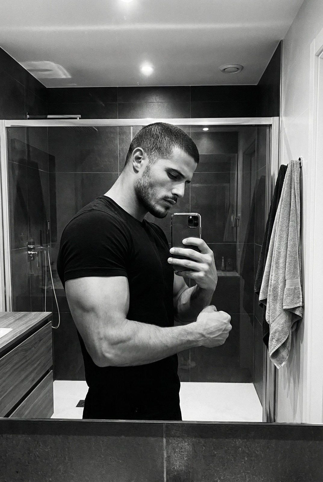 A muscular man taking a black and white mirror selfie in a modern bathroom with dark walls and a glass shower.