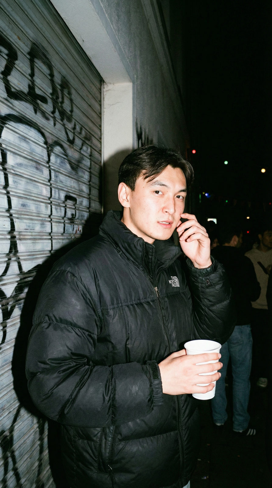 A gritty flash-photography portrait of a person at a house party, wearing an oversized puffer jacket, against a metal shutter wall.