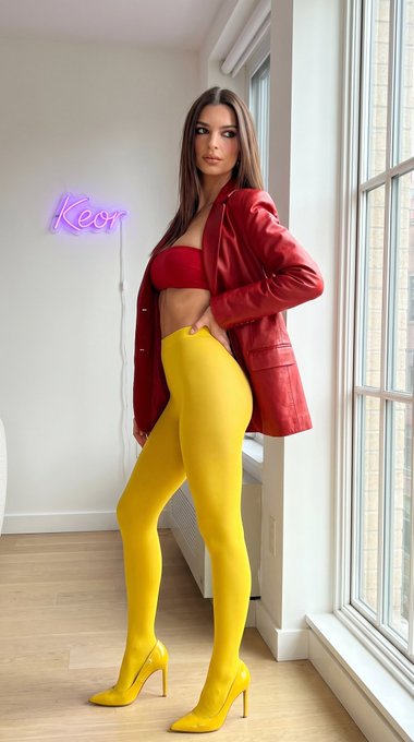 Full-body shot of a woman in a red blazer and yellow tights, posed against a neon sign and cityscape.