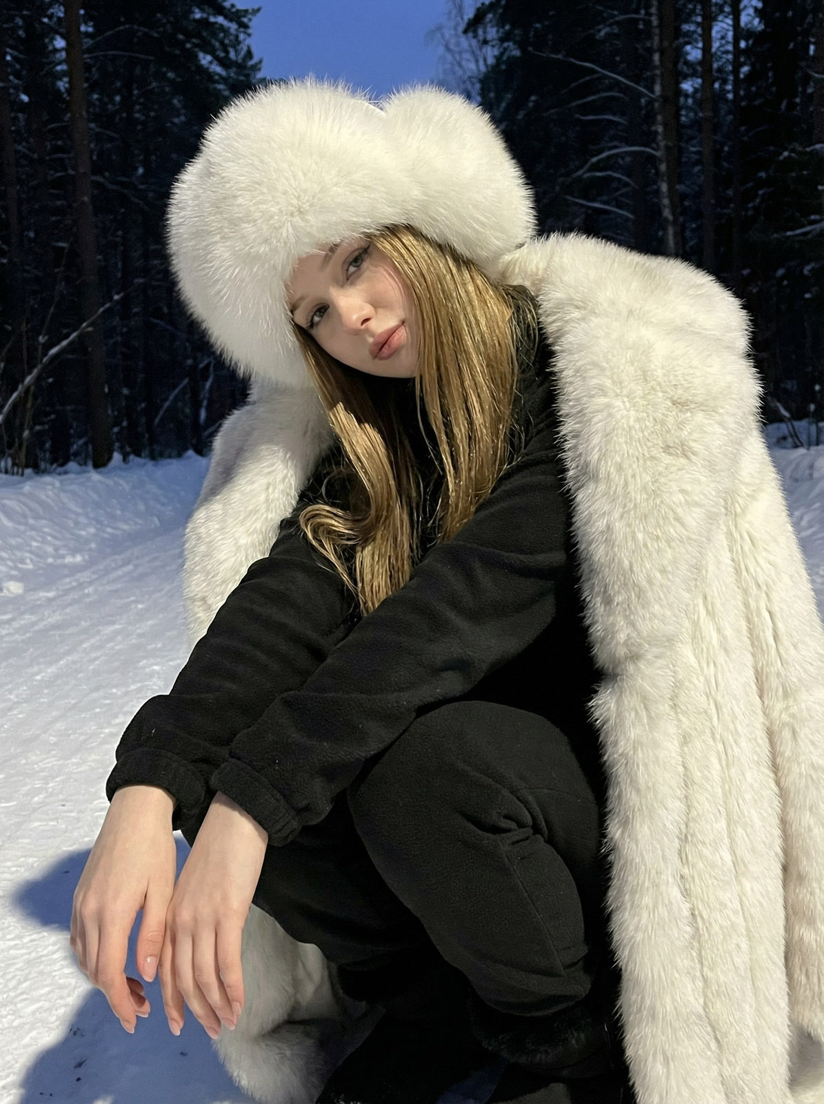 Close-up of a woman in a snowy forest, wearing a fur coat and hat, sitting relaxed with her knees bent.
